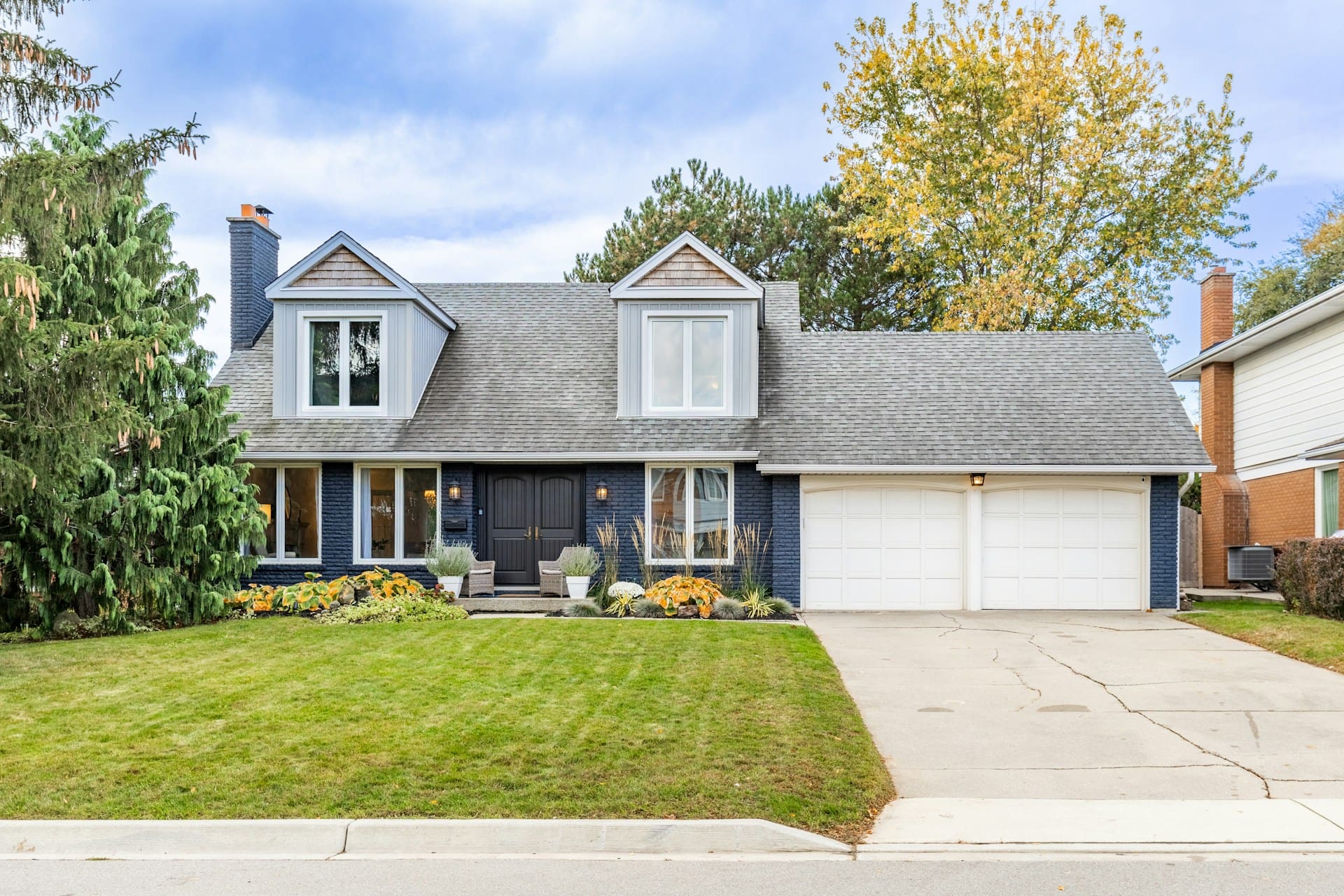 A two-story house with a dark brick facade, double garage, front porch with wicker chairs, and a well-maintained lawn with colorful autumn plants under a partly cloudy sky.