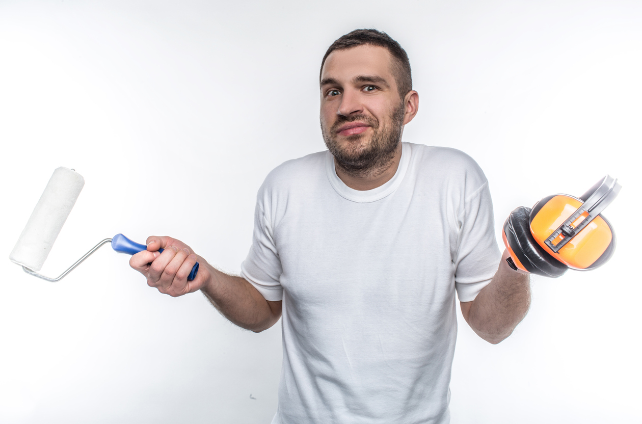 A do-it-yourself house painter holding a brush roller and an ear protector standing against a white wall.