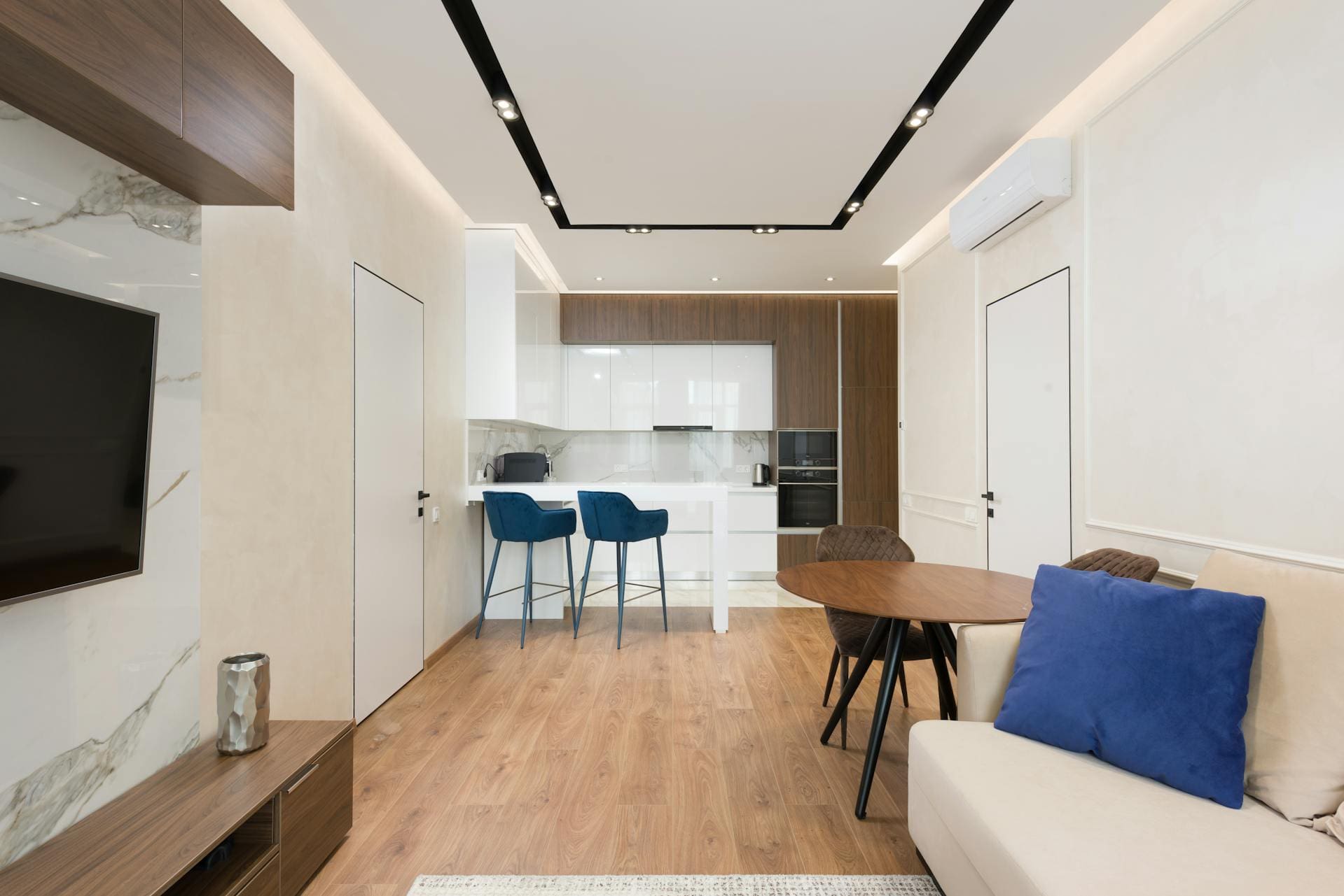 A modern living room with a wooden floor, white and wood kitchen with blue bar stools, a round dining table, a beige sofa with a blue cushion, and a TV on a marble wall, illuminated by recessed ceiling lights.