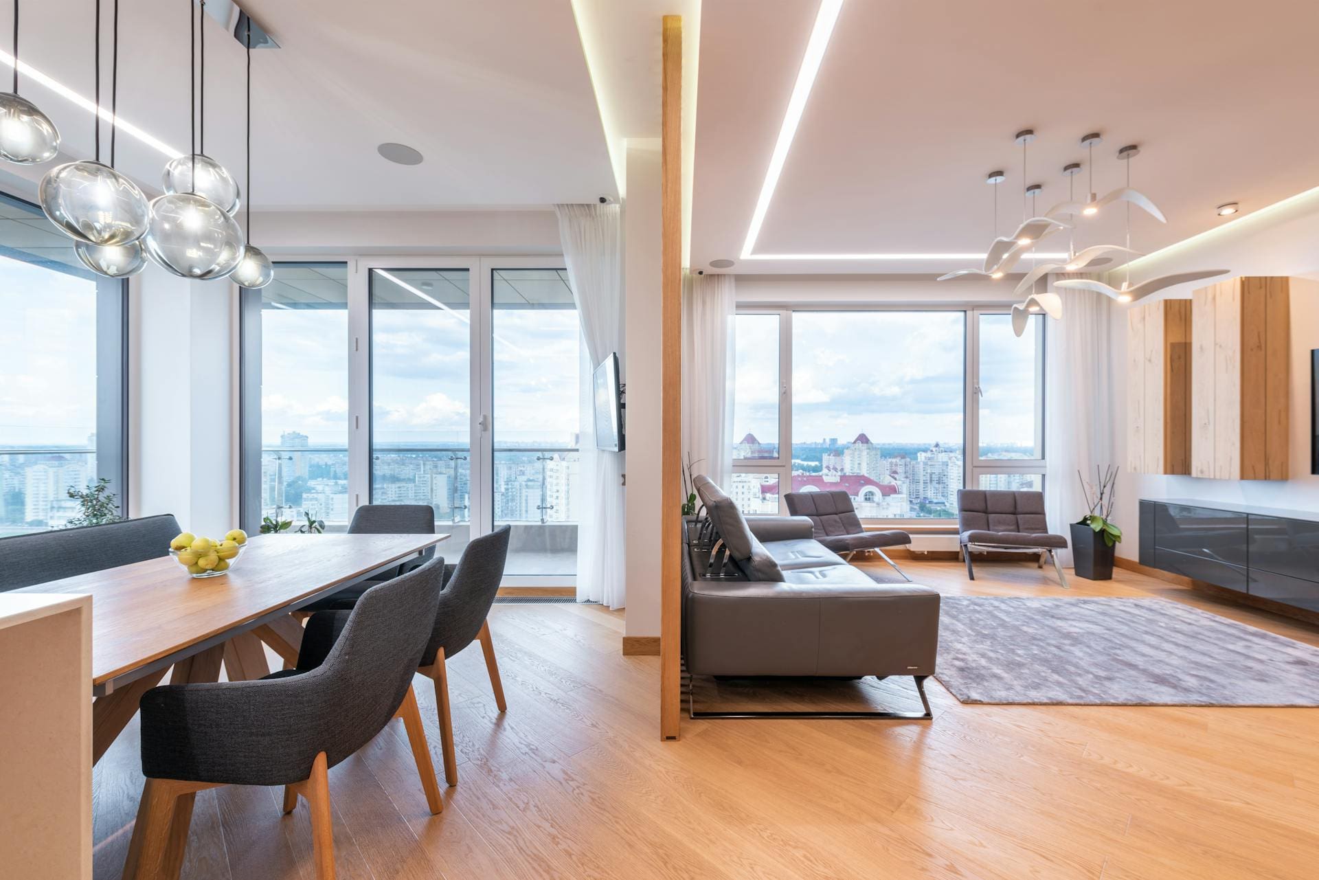 A modern open-plan living and dining area with wooden flooring, a long dining table with chairs and fruit bowl, a gray sofa, and large windows offering a city view, illuminated by stylish pendant lights.