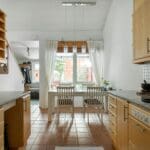 A bright kitchen with wooden cabinets, a granite countertop, a dining table with two chairs, and large windows with curtains, leading to a staircase.