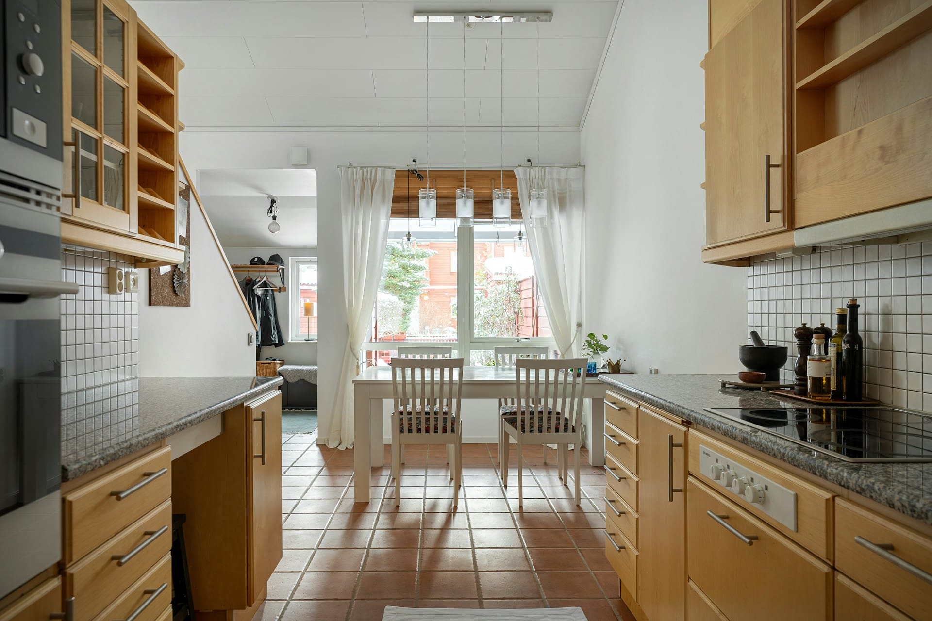 A bright kitchen with wooden cabinets, a granite countertop, a dining table with two chairs, and large windows with curtains, leading to a staircase.