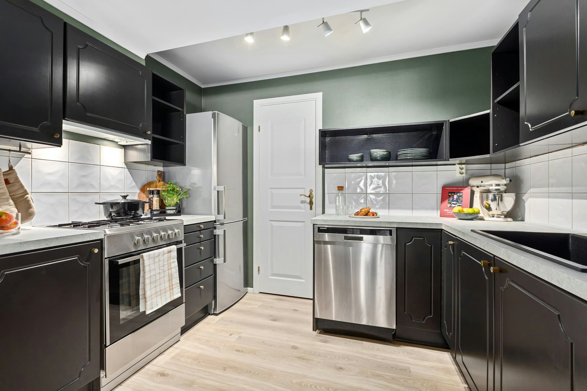 A modern kitchen with black cabinets, white countertops, a stainless steel oven and dishwasher, a refrigerator, and wooden flooring, illuminated by ceiling spotlights.