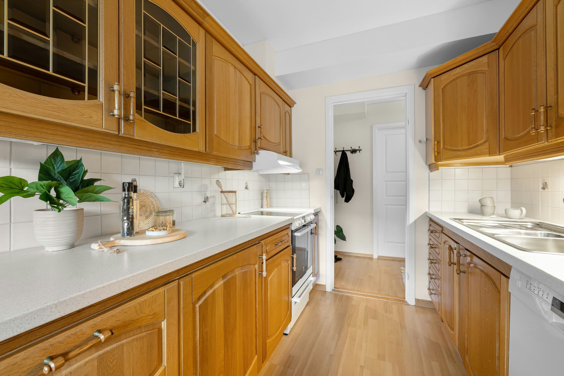 A cozy kitchen with wooden cabinets, white countertops, a sink, oven, and potted plants, featuring a doorway to another room with wooden flooring.