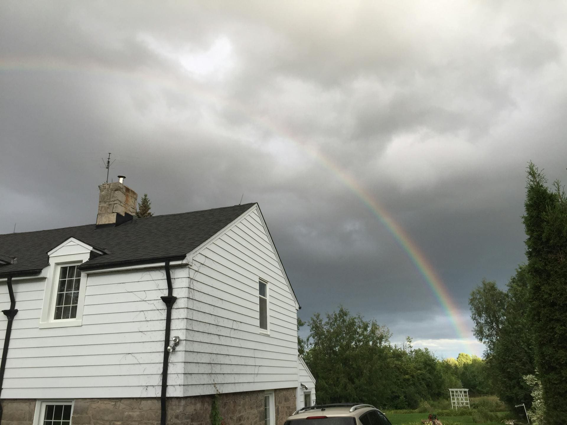 A white house with a dark roof under a cloudy sky, featuring a rainbow arching over it, with trees and a car in the foreground.