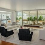 A modern open-plan living room with white sofas, black armchairs, a glass coffee table, and large sliding doors opening to a lush outdoor patio with plants and seating.