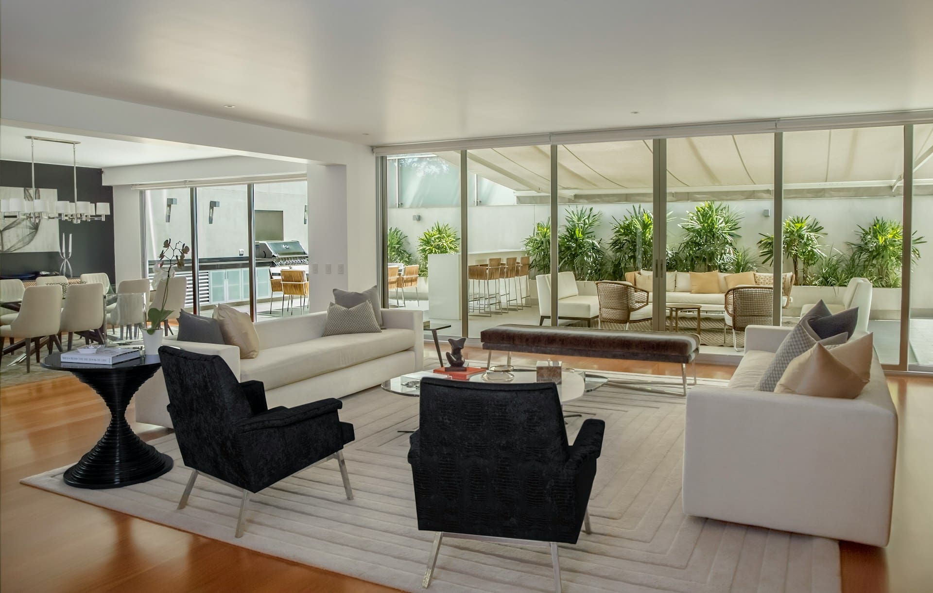 A modern open-plan living room with white sofas, black armchairs, a glass coffee table, and large sliding doors opening to a lush outdoor patio with plants and seating.