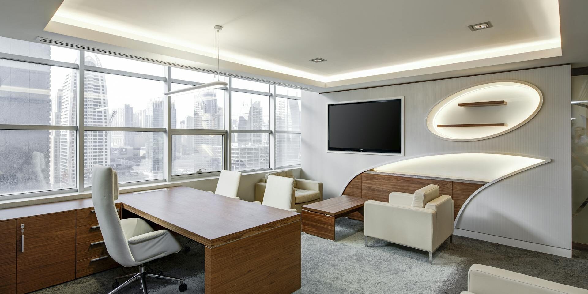 A modern office with a wooden desk, white ergonomic chair, beige sofas, a large TV, and large windows offering a city skyline view, featuring ambient ceiling lighting