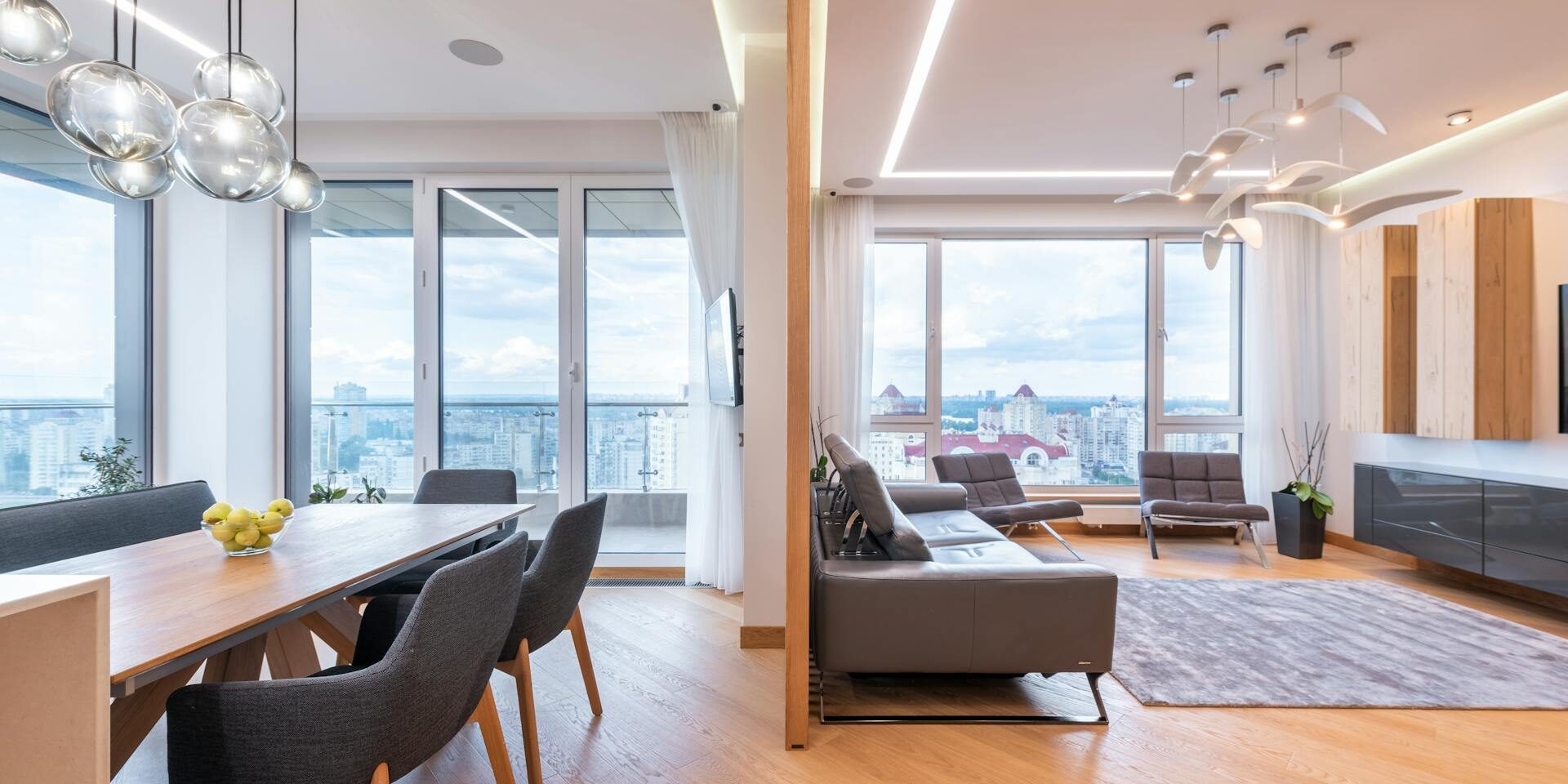 A modern open-plan living and dining area with wooden flooring, a long dining table with chairs and fruit bowl, a gray sofa, and large windows offering a city view, illuminated by stylish pendant lights.