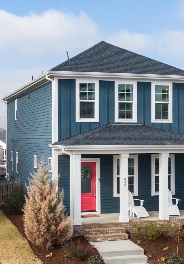 A blue house with a red door and white trim, featuring a small porch with chairs, surrounded by a fenced yard and neighboring houses, under a partly cloudy sky.