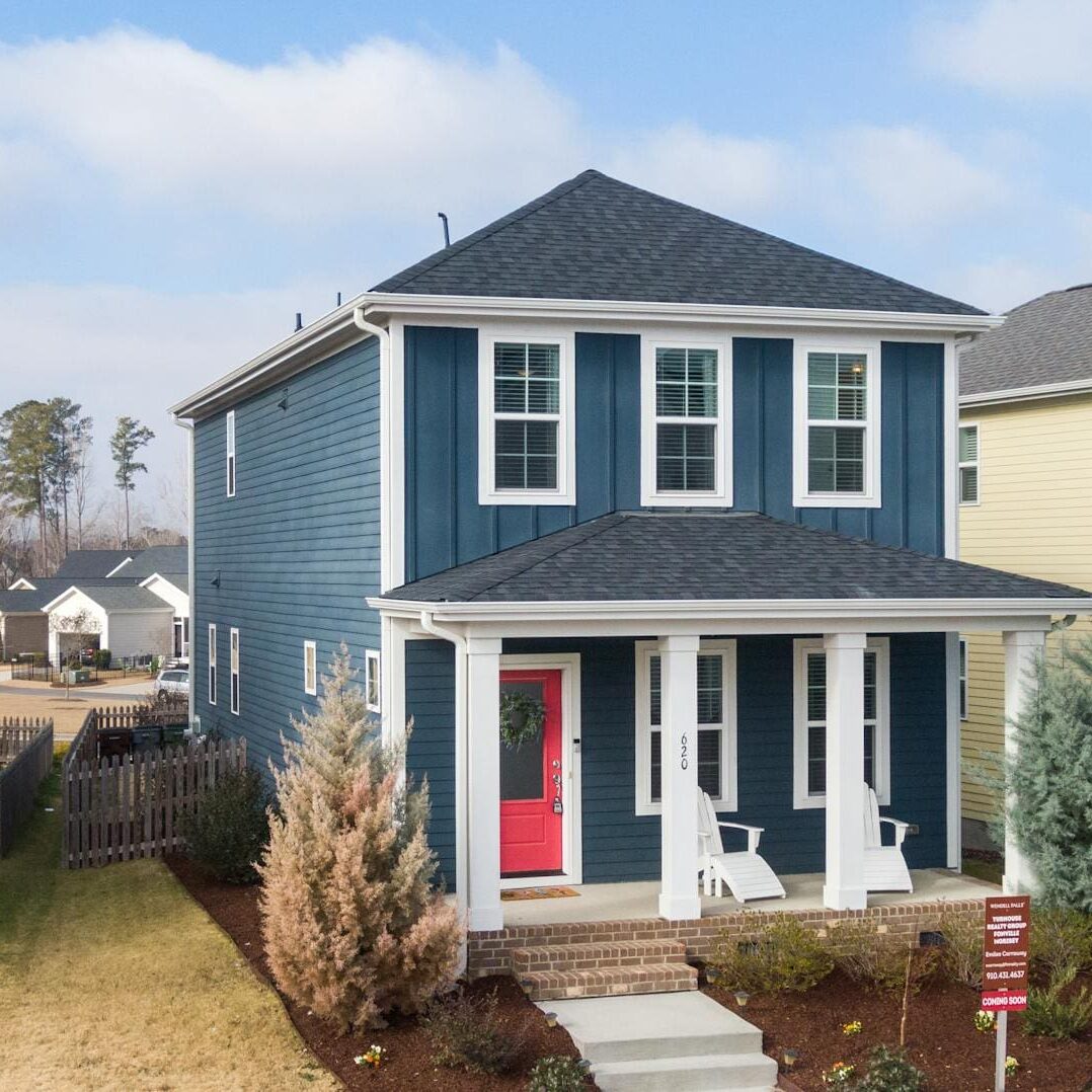 A blue house with a red door and white trim, featuring a small porch with chairs, surrounded by a fenced yard and neighboring houses, under a partly cloudy sky.