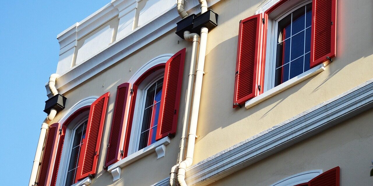 Classic building facade with pale yellow walls, red shutters, and white trim under a clear blue sky, exuding a charming and timeless vibe.