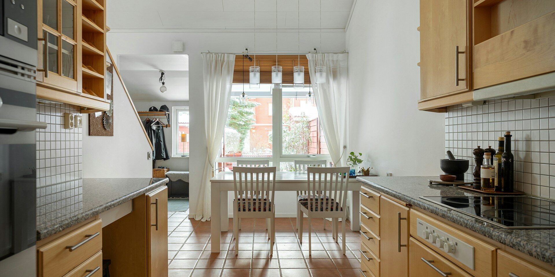 A bright kitchen with wooden cabinets, a granite countertop, a dining table with two chairs, and large windows with curtains, leading to a staircase.