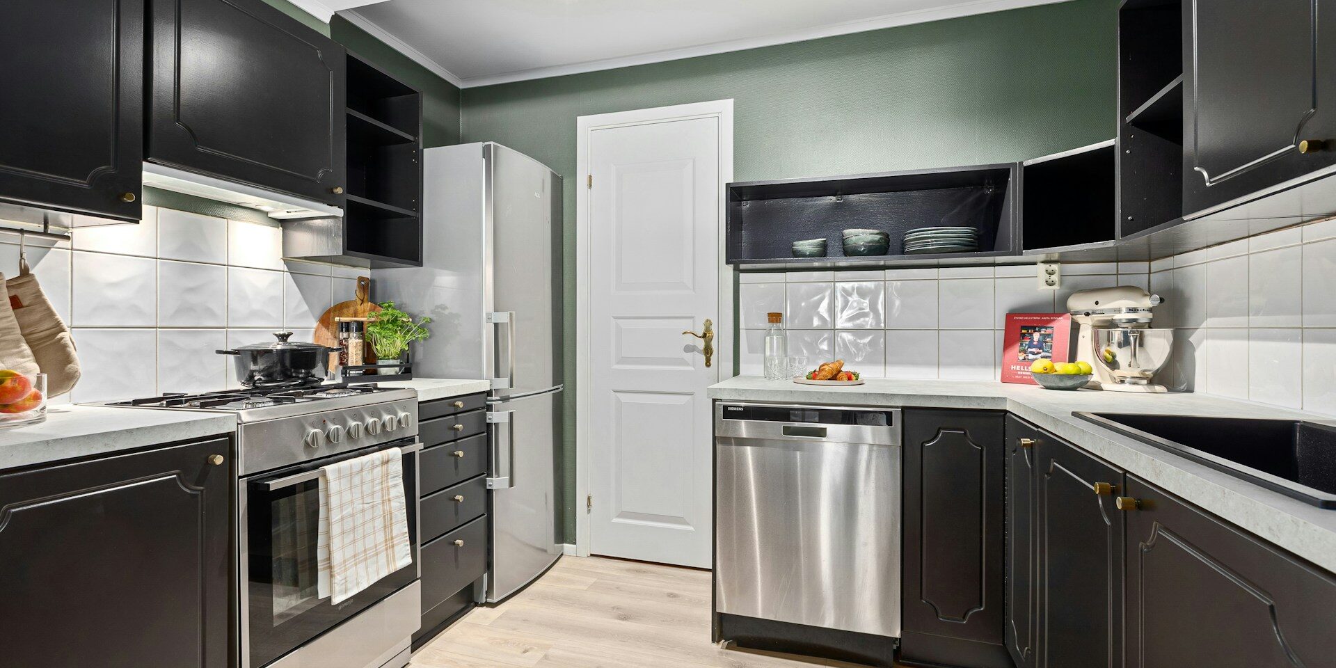 A modern kitchen with black cabinets, white countertops, a stainless steel oven and dishwasher, a refrigerator, and wooden flooring, illuminated by ceiling spotlights.