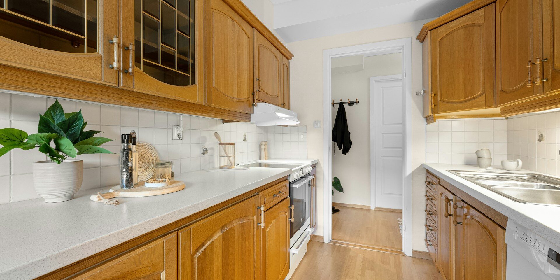 northern-virginia-cabinet-painting-services (3) A cozy kitchen with wooden cabinets, white countertops, a sink, oven, and potted plants, featuring a doorway to another room with wooden flooring.