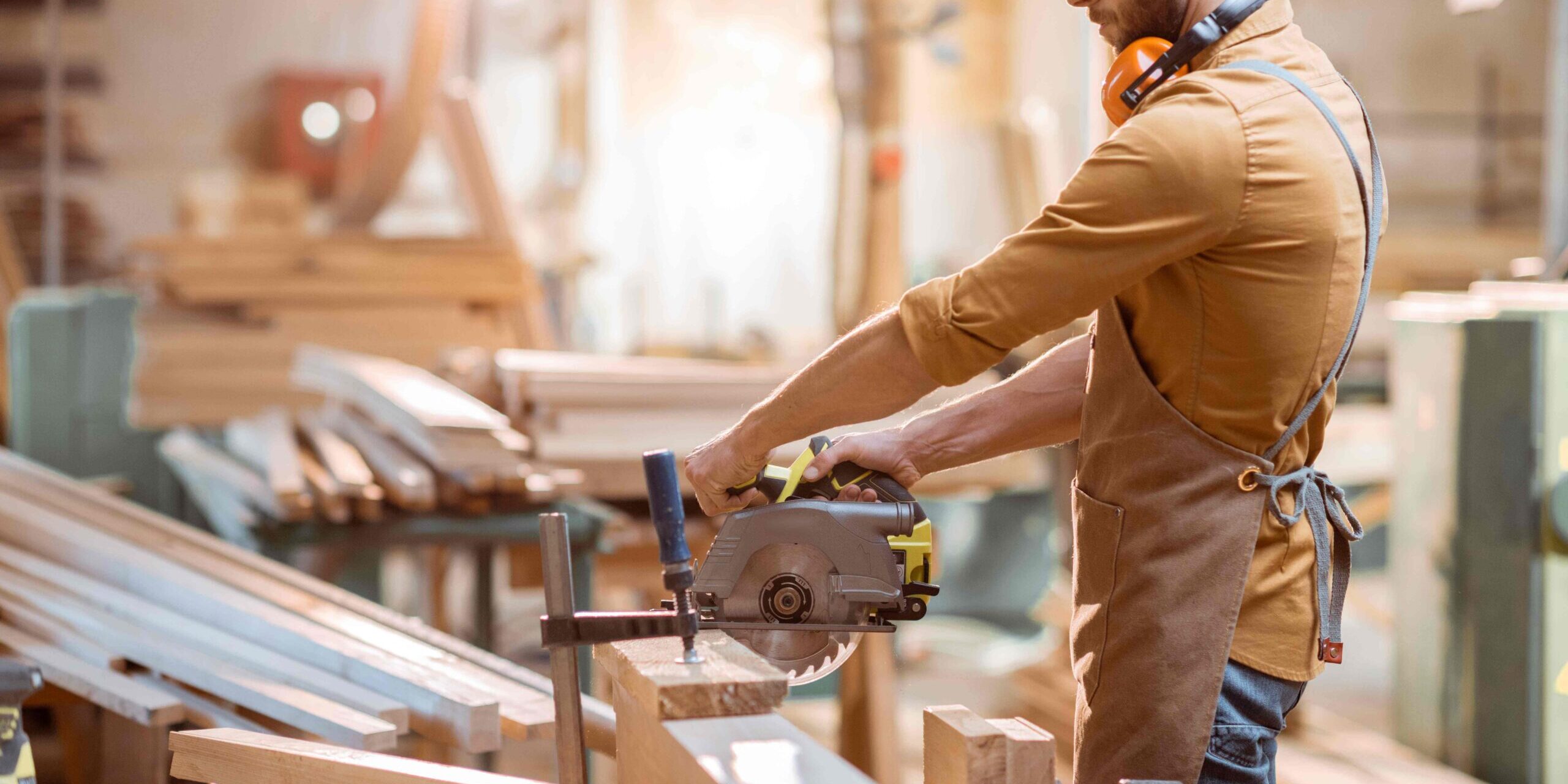 A person wearing an apron and ear protection using a circular saw to cut wood in a workshop filled with tools and lumber.