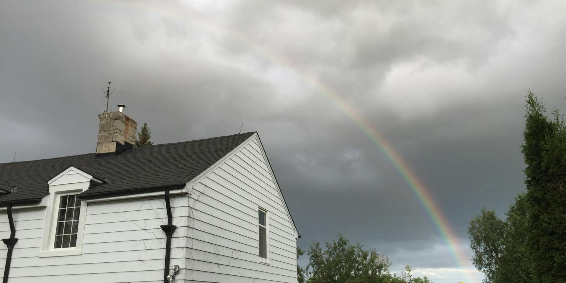 A white house with a dark roof under a cloudy sky, featuring a rainbow arching over it, with trees and a car in the foreground.