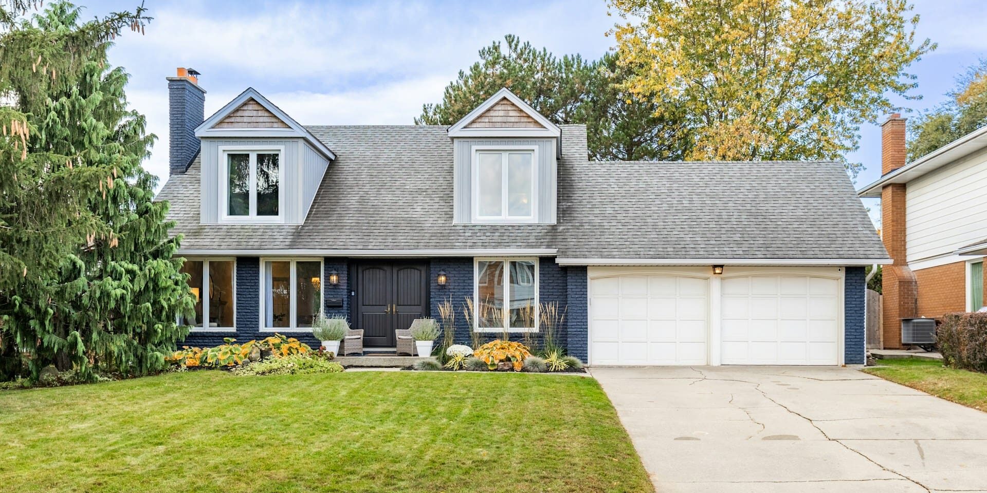A two-story house with a dark brick facade, double garage, front porch with wicker chairs, and a well-maintained lawn with colorful autumn plants under a partly cloudy sky.