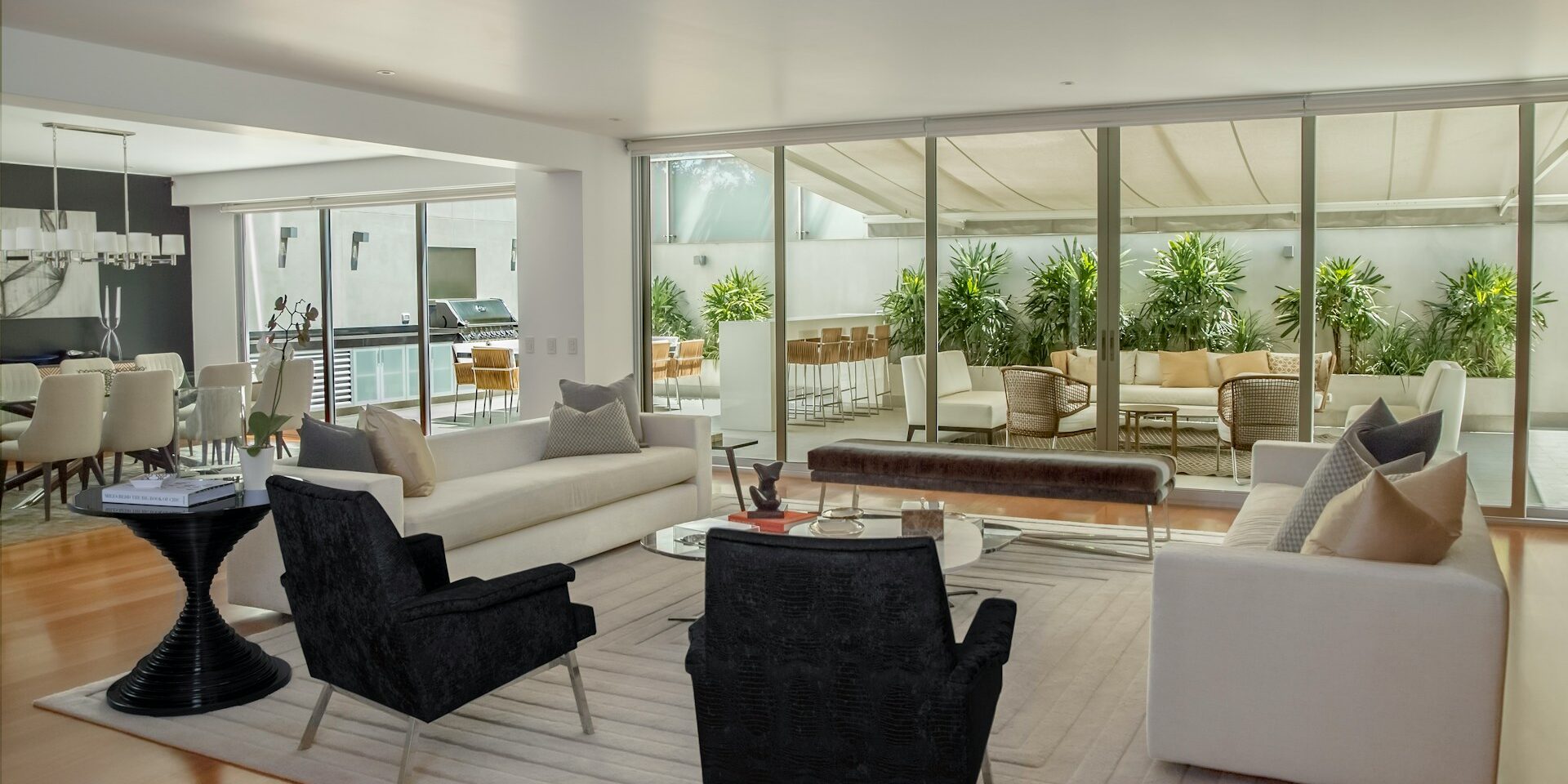 A modern open-plan living room with white sofas, black armchairs, a glass coffee table, and large sliding doors opening to a lush outdoor patio with plants and seating.
