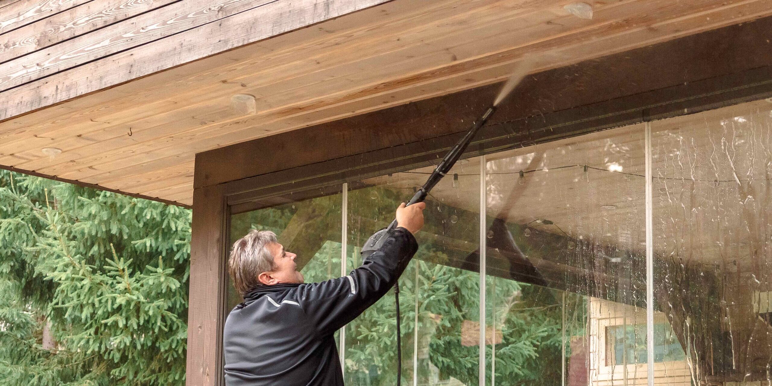 A person in a black jacket using a pressure washer to clean the wooden ceiling and glass walls of a covered outdoor area, with trees visible in the background.
