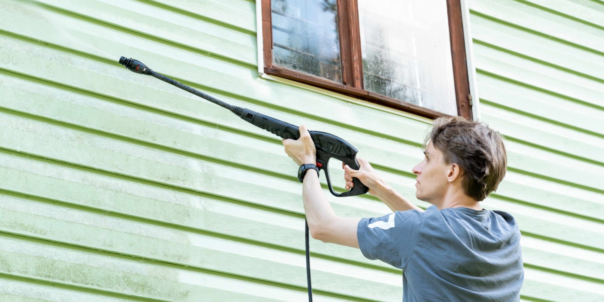 A person in a gray shirt using a pressure washer to clean the green siding of a house near a window.